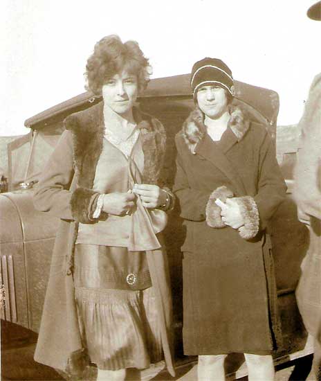 <glamor girls eddie may rombold pearl downer wearing fur trimmed coats, standing in front of old cars model a model t>