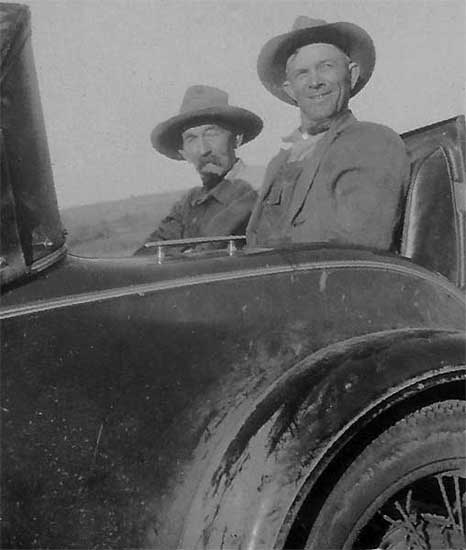 <bruce downer, along with an old character, sitting in the rumble seat of an old car. spoke wheels. old character with a moustache and floppy hat>