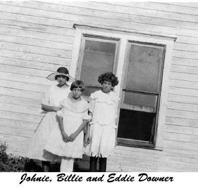 <three downer girls Johnie, wearing a large brimmed hat, Billie and Eddie posed by a house with open windows>