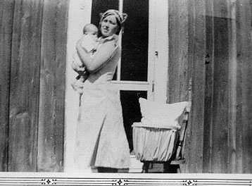 <johnie holding baby adrienne by east door of ramon ranch house. baby bassinet sitting on oak dinette chair lock can seen hanging on a nail on the wall of the house>