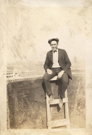 <Man on wooden ladder sitting on the edge of a steel tank. May be a water tower tank. >