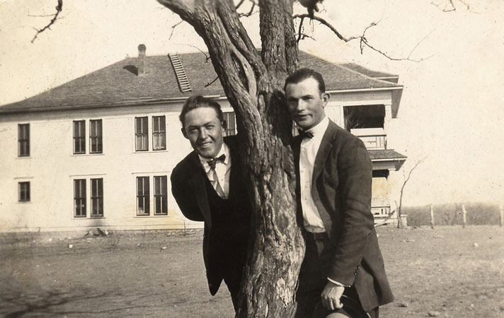 <two men standing by tree. One man peaking around the tree. two story building in background. both men wearing bow ties.>