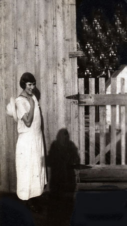 <Lady standing by building and gate with hat draped over her shoulder restored by terry>