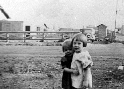 <young adrienne and donna rombold hugging houses fence and old car and outdoor toilet in background>