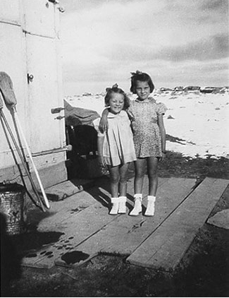 <sheila and donna rombold standing on planks which serves as a porch. Snow on the ground. Broom and mop bucket shown in the photograph>
